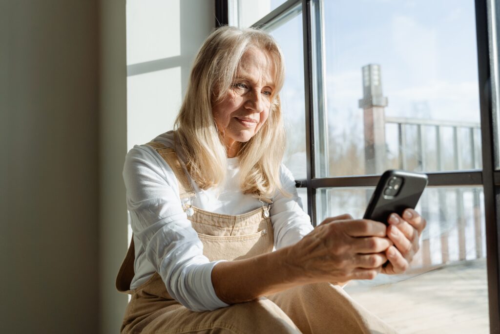 Seniorin sitzt am Fenster mit Handy in der Hand