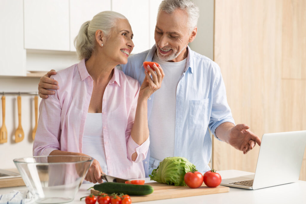 Picture of cheerful mature loving couple family standing at the kitchen using laptop computer and cooking. Looking aside.